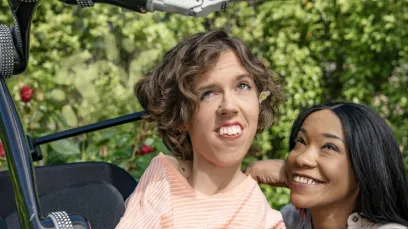 A white person with wavy brown hair sits in their motorized wheelchair with a robot arm attachment holding an Android phone. A Black woman crouches next to them and smiles up at the Android phone.