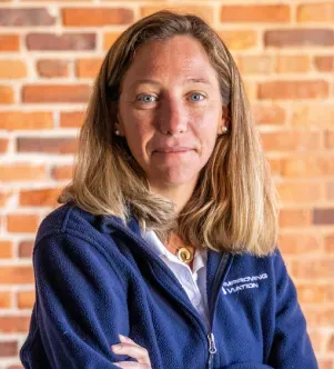 A woman with her arms crossed wearing a navy blue fleece jacket with "Improving Aviation" printed on the chest, standing in front of a brick wall.