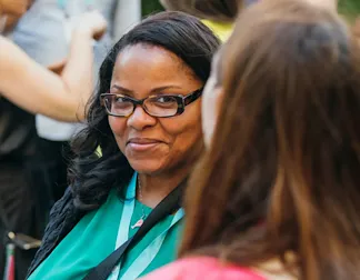 A dark woman with long hair and wearing spectacles smiling at other woman in crowd