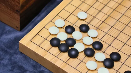 A high-angle, close-up shot of a wooden Go board (goban) featuring several black and white stones placed during a match. A traditional wooden bowl for holding stones is visible in the upper-left corner, set against a dark blue textured tablecloth. The image symbolizes the AlphaGo AI breakthrough in strategic gameplay.