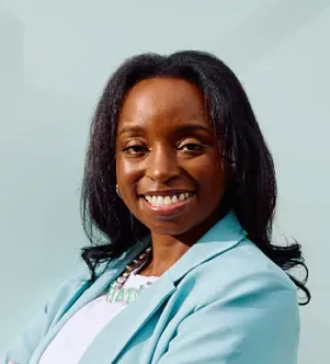 A smiling Black woman with dark shoulder-length hair wearing a light blue blazer and a silver necklace, posing confidently against a pale blue background.