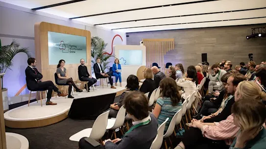A wide-angle view of a professional panel discussion on a stage titled "Fighting Misinformation Online." Five panelists are seated on a raised platform facing a seated audience. The room features modern decor with light wood accents and potted palm plants.