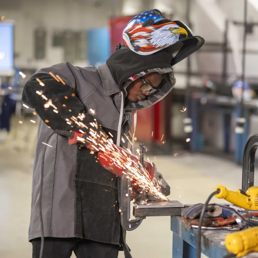 A young person wearing a welding helmet with an American flag and eagle design, protective glasses, and a grey jacket with leather sleeves is using an angle grinder on a metal piece at a workbench, creating a shower of bright orange sparks.