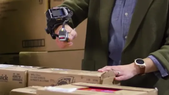 A person using a wearable finger barcode scanner to scan cardboard boxes in a warehouse or pharmacy setting.