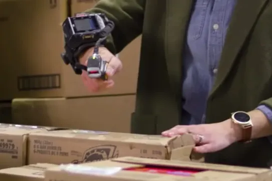 A person using a wearable finger barcode scanner to scan cardboard boxes in a warehouse or pharmacy setting.