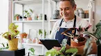 A smiling man wearing a denim apron holds a potted plant and looks at a digital tablet inside a brightly lit plant shop.