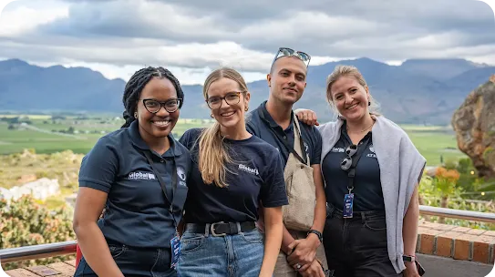 Four young adults wearing matching navy blue "Global Changemakers" shirts and event lanyards stand close together and smile for a photo. They are outdoors on a brick overlook with a vast green valley and layered mountain range under a cloudy sky behind them.