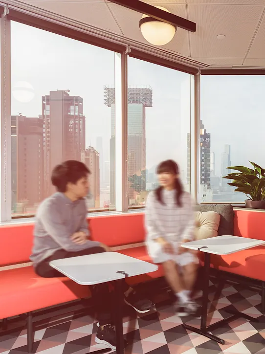 Two people sit on a red couch in front of windows.