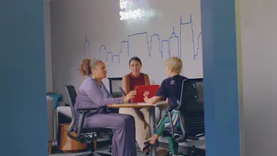 three women sitting around a table with a laptop talking.