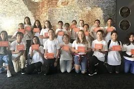 A large group of diverse middle school students smile and pose while holding orange Thimble.io STEM kits in front of a mosaic wall.