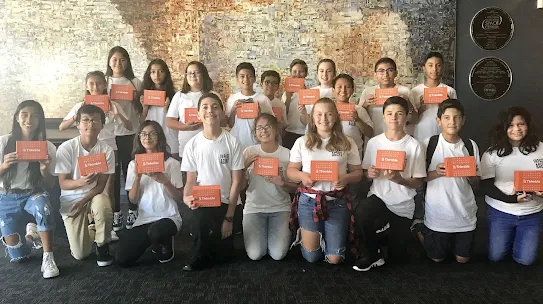 A large group of diverse middle school students smile and pose while holding orange Thimble.io STEM kits in front of a mosaic wall.