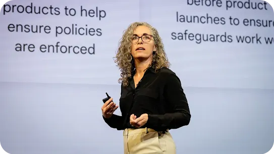 A woman with curly grey hair and glasses wearing a black blouse gives a presentation on stage. Behind her, a large screen displays text about "products to help ensure policies are enforced" and "safeguards work." She is holding a slide clicker and speaking.
