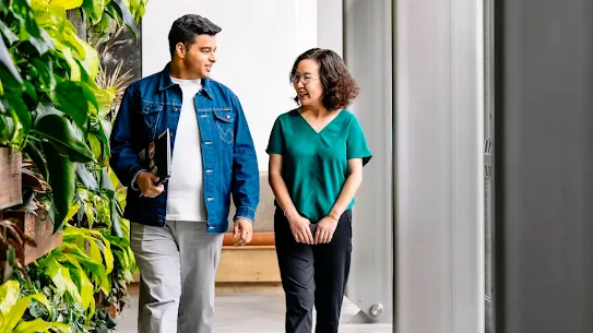 Two colleagues walk side-by-side down a bright, high-ceilinged office hallway, engaged in a friendly conversation. On the left, a man wearing a denim jacket over a white t-shirt and grey trousers carries a laptop and looks toward his companion. Beside him, a woman with short dark hair, wearing a green V-neck blouse and black trousers, smiles back as they walk. To their left, a vibrant vertical garden wall overflowing with lush green plants like Monstera and neon pothos adds a natural element to the space. Large, minimalist grey pillars line the right side of the corridor, enhancing the modern, industrial feel of the workplace.