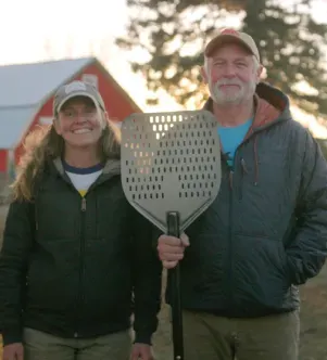 A portrait of a woman and man standing outdoors on a farm in South Dakota at sunset; the man holds a large metal pizza peel, and a red barn is visible in the background.