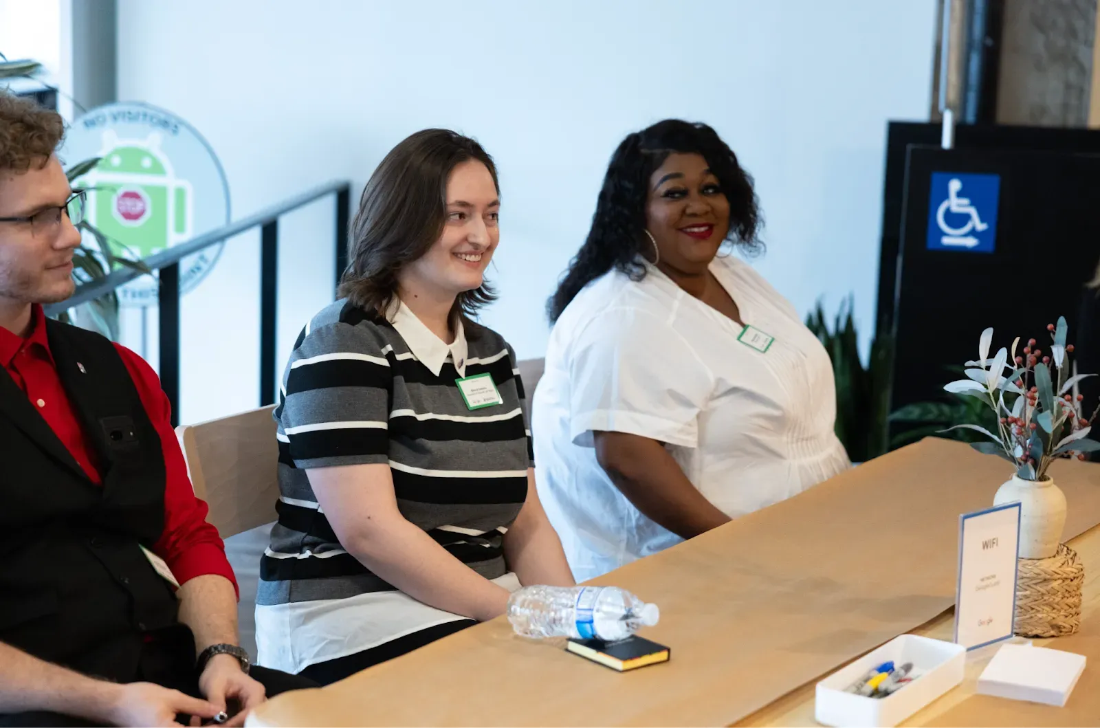 Bianca Ionescu, wearing a black and white striped shirt, and Hannah Brown, in a white blouse, sit at a long table during a Google cybersecurity event. They are both smiling and looking forward. A small plant and a "WiFi" sign are on the table in front of them.