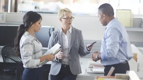 A medium shot of three colleagues in professional business attire, including a woman holding a phone, as they stand and talk during a brief impromptu meeting.
