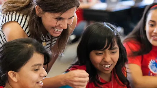 A smiling female teacher leans over and points at a laptop screen, engaging with three young female students who are also smiling in a classroom.