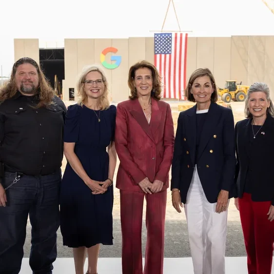 A group of six people stand smiling on a white platform. They are outdoors with a large data center featuring a Google logo in the background.