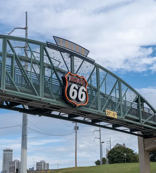 A green metal pedestrian bridge in Tulsa, Oklahoma, featuring a large neon Route 66 sign and the city name, set against a cloudy blue sky.