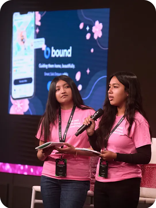 Two girls in pink shirts presenting on stage with a microphone; a screen behind them displays a presentation for an app called "bound".