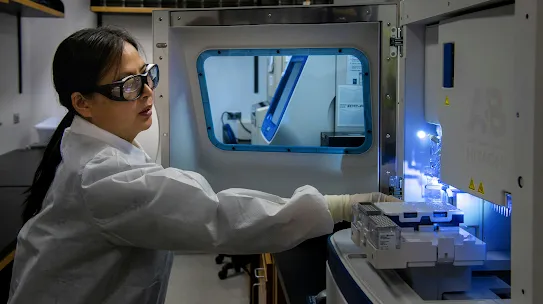 A photograph of a medical researcher operating advanced laboratory equipment, used to illustrate the Department of Defense (DoD) case study on battling cancer. The image represents the intersection of medical science and AI technology, highlighting efforts to improve early detection and treatment outcomes for service members and the public.