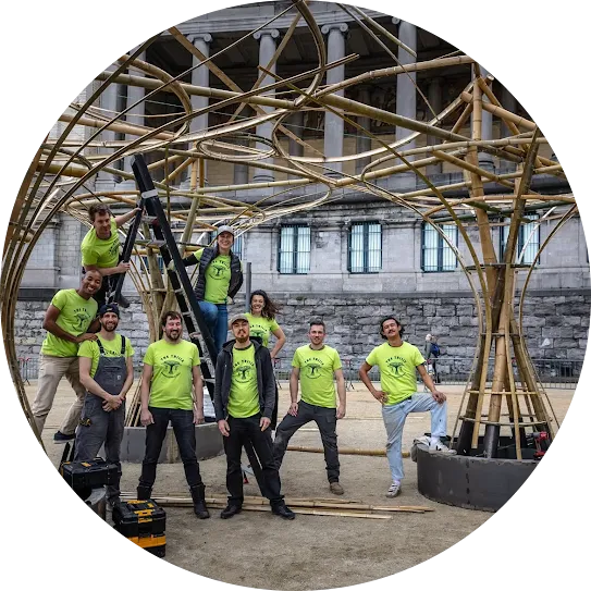 A group of eight construction team members in matching neon-green "The Trilix" t-shirts smiling and posing inside a large, intricate, hand-crafted bamboo architectural pavilion structure outdoors.